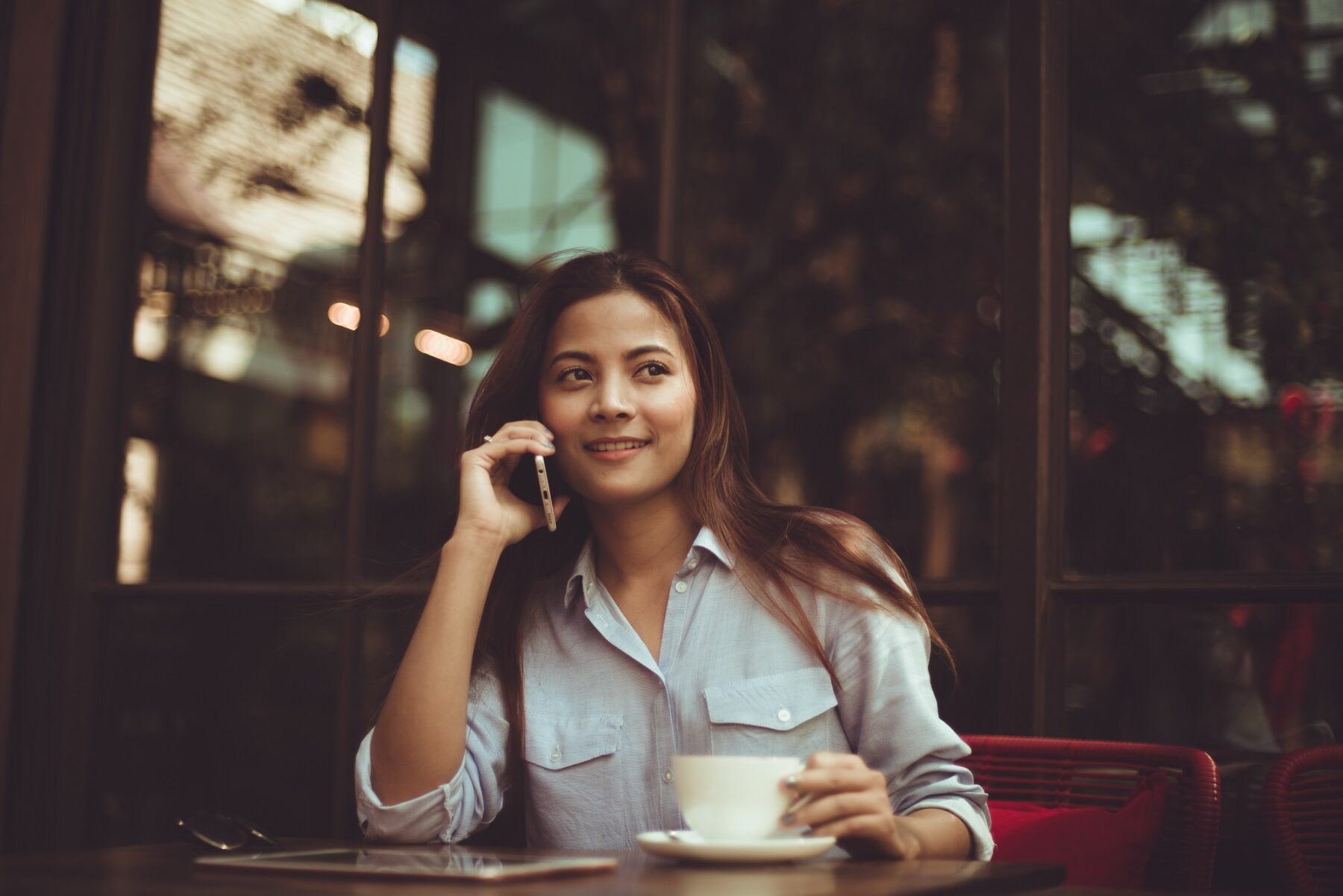 A woman sits at a cafe table, smiling while talking on her phone and holding a cup of coffee. She wears a light blue shirt and has long brown hair, with a tablet on the table in front of her.